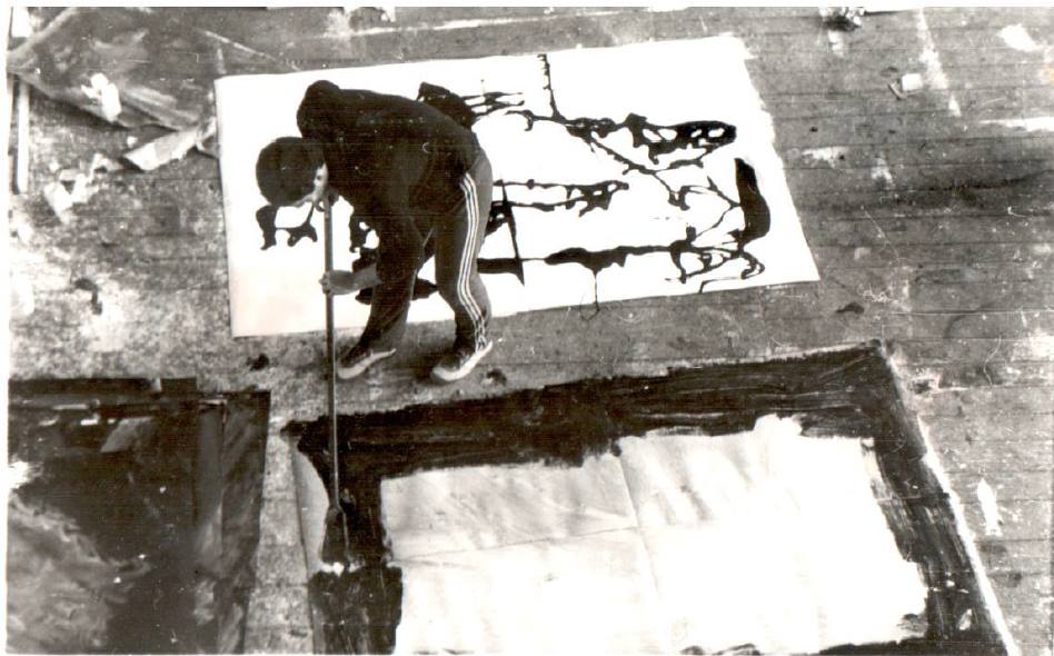 Black and white photograph showing a person bending over a large canvas on a wooden floor, applying paint with a long brush or roller. The scene includes abstract artwork with bold, dark lines and shapes on the canvases, highlighting an artistic process in progress.