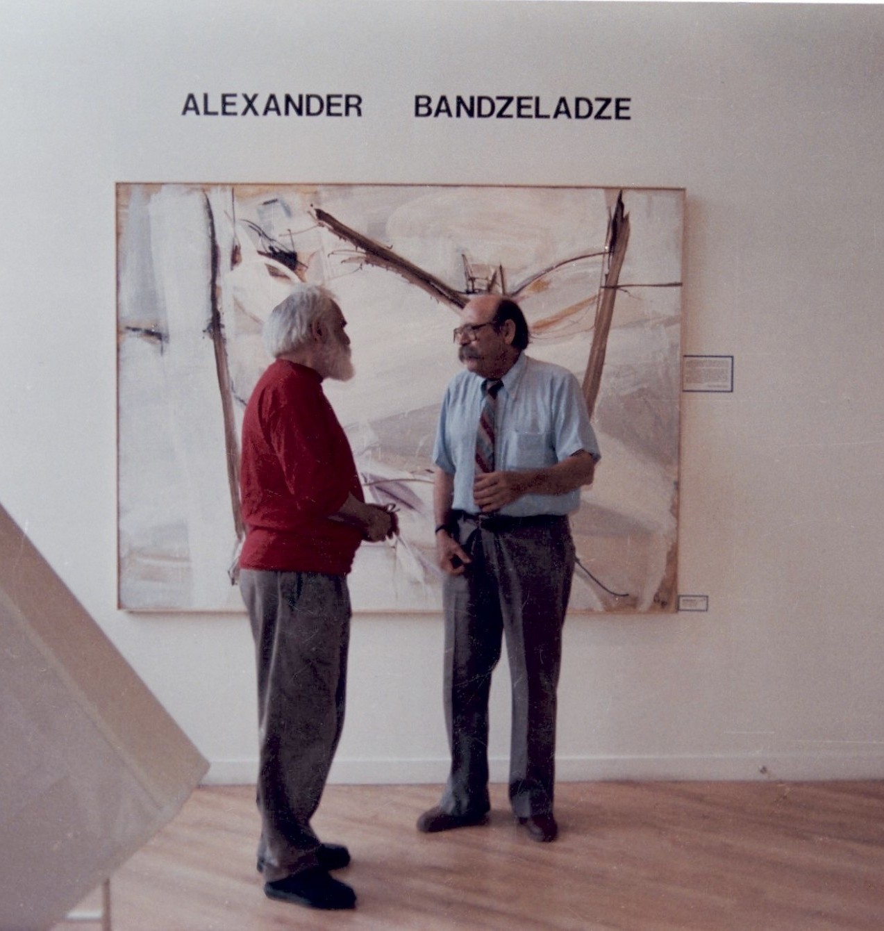 Photograph of two men engaged in conversation inside an art gallery, standing in front of a large abstract painting with bold brushstrokes and muted colors. Text above the painting reads "ALEXANDER BANDZELADZE," indicating the artist's name and highlighting the gallery's focus on his work.