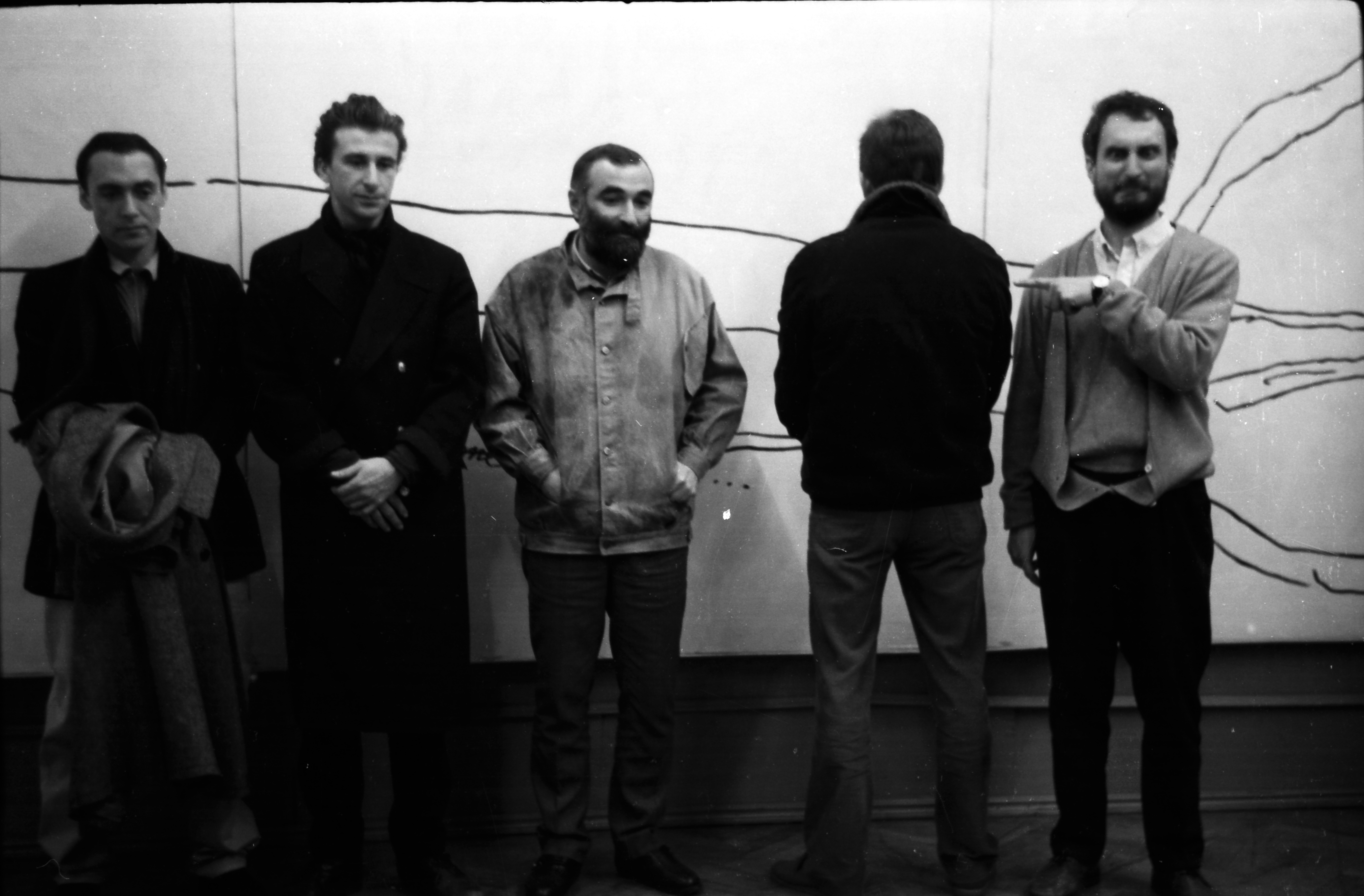 Black and white photograph showing five men standing in front of a wall with abstract line drawings. Four men face forward with blurred faces, while one man faces away, highlighting varied postures and casual attire.
