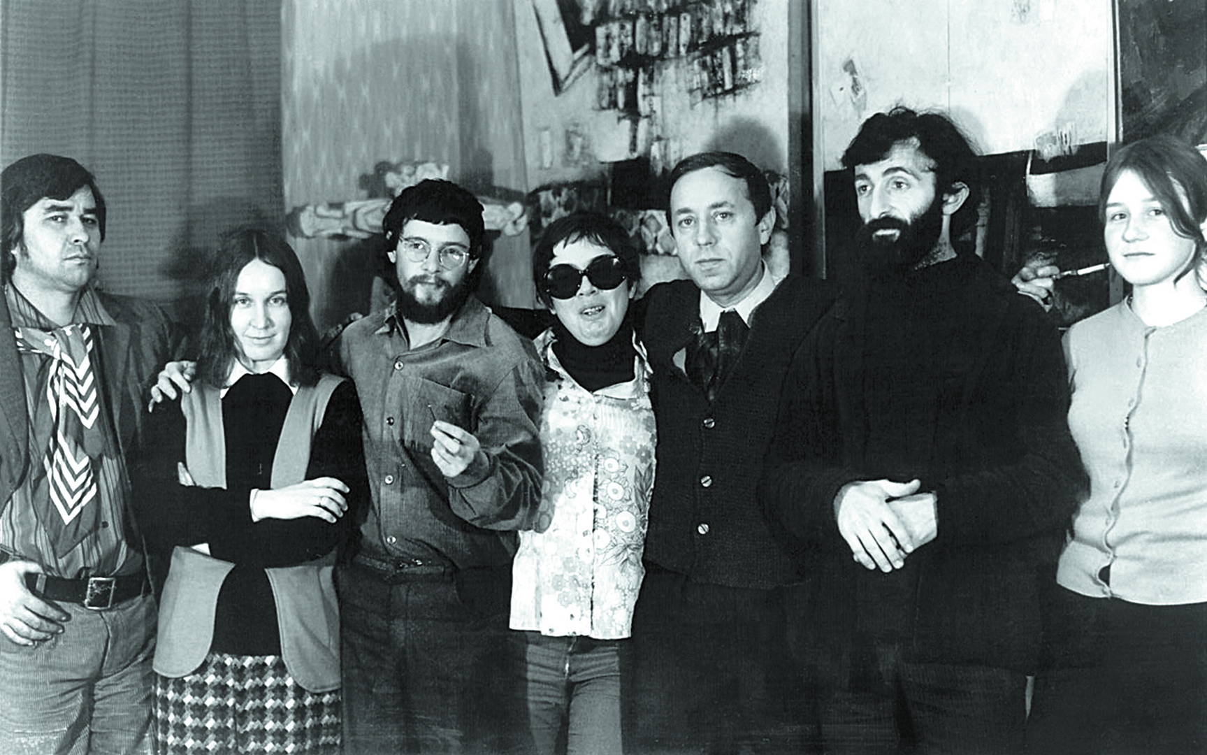Black and white photograph showing a group of eight people standing closely together indoors, with faces blurred for anonymity. The setting appears casual with varied clothing styles, and the background includes wall decorations and framed pictures.