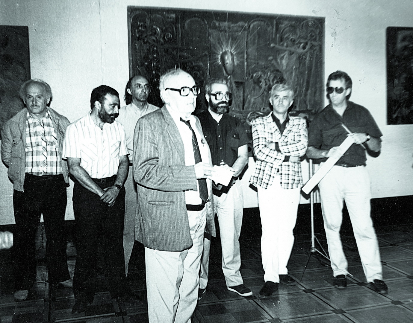 Black and white photograph showing seven men standing indoors, with one man in the foreground holding papers and speaking. Background features a large decorative wall panel, and men wear a mix of casual and semi-formal attire, suggesting a formal or professional gathering.