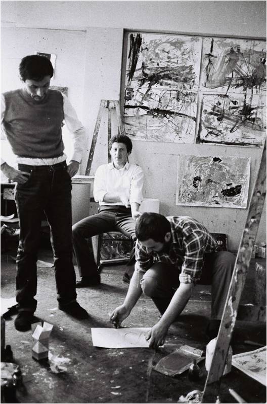 Black and white photograph showing three men in an art studio with abstract paintings on walls and scattered art supplies on floor. One man is crouched drawing on paper, another sits on a chair, and the third stands with hands on hips, suggesting a collaborative creative process.