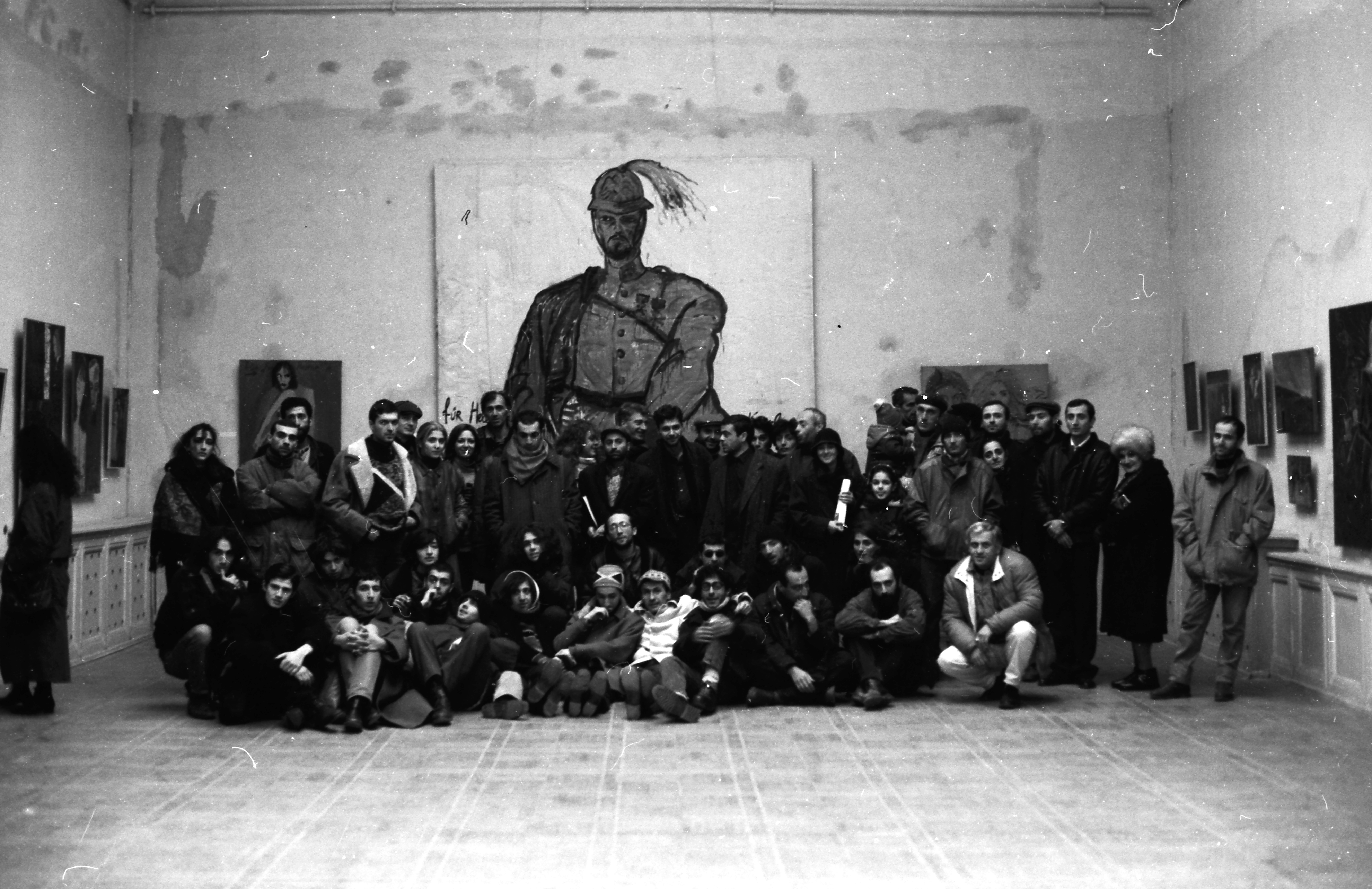 Black and white photograph of a large group of people posing inside an art gallery with paintings displayed on walls. Central focus is a prominent large painting of a figure wearing a hat, surrounded by smaller artworks and diverse individuals in casual attire.