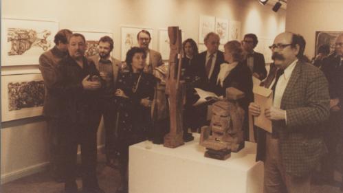 Photograph of a group of people gathered in an art gallery, observing and discussing wooden sculptures displayed on a white pedestal. The setting includes framed artwork on walls, warm lighting, and individuals holding papers or folders, indicating an art exhibition or critique event.