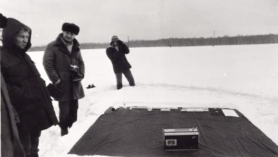 Black and white photograph showing three people standing near a large black cloth spread on snow-covered ground with a box placed in the center. 
