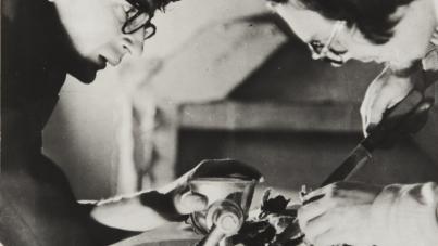 Black and white photograph showing two individuals working together on a manual task involving a grinder and a knife. One person holds a grinder while the other cuts newspaper, highlighting a collaborative effort in a workshop setting.