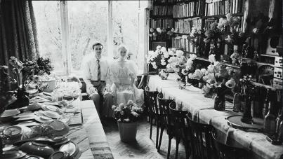 A black and white photo of two people sitting by a window and two tables lined with flowers.