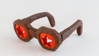 Photograph of a pair of novelty glasses with thick, brown, angular frames and large, red, star-shaped lenses. The glasses have a blocky, handcrafted appearance and are displayed against a plain white background.