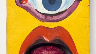A photograph of a colorful wooden sculpture featuring a large blue eye above an open mouth with red lips and a protruding tongue, set against a bright yellow rectangular background. The sculpture combines bold colors and exaggerated facial features to create a surreal and expressive visual effect.