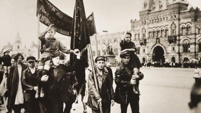 Black and white photograph depicting a group of people marching in a public square, carrying flags and children on their shoulders. The background features historic architecture, suggesting a significant political or social demonstration in an urban setting.