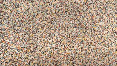 Photograph of a densely packed abstract artwork featuring numerous small, colorful symbols and shapes scattered across a light background. The composition includes varied hues of orange, blue, green, and black, creating a vibrant, chaotic pattern without a clear focal point or recognizable figures.