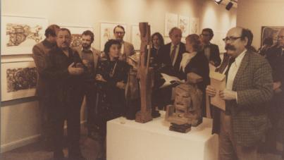 Photograph of a group of people gathered in an art gallery, observing and discussing wooden sculptures displayed on a white pedestal. The setting includes framed artwork on walls, warm lighting, and individuals holding papers or folders, indicating an art exhibition or critique event.