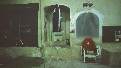 Photograph of a dimly lit, worn indoor space featuring a hanging fish in an arched alcove and a white chair draped with a red cloth marked by a white cross. Surrounding elements include a dark rectangular panel on the left, a small display on the right, and various wires and objects scattered on a tiled floor, evoking a mysterious or artistic atmosphere.