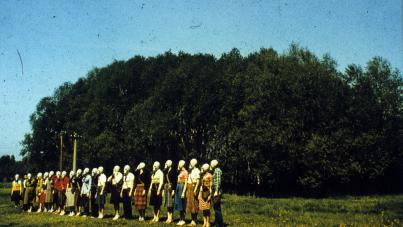 Photograph showing a group of people standing in a straight line outdoors on grassy field with dense trees in background. Individuals wear casual clothing and white gas masks.