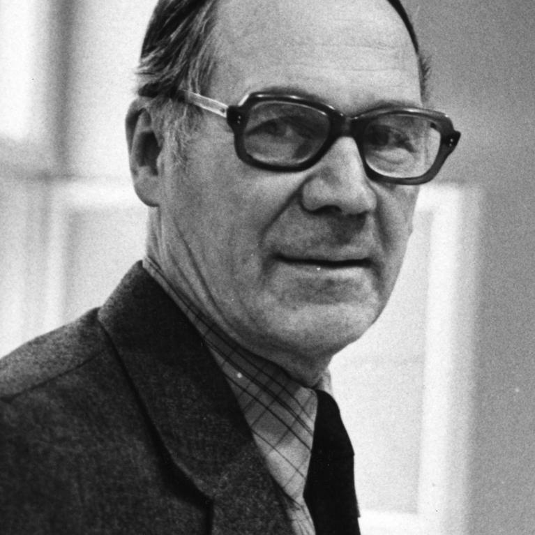 Headshot of the artist Evald Okas wearing thick-rimmed glasses and a black suit.