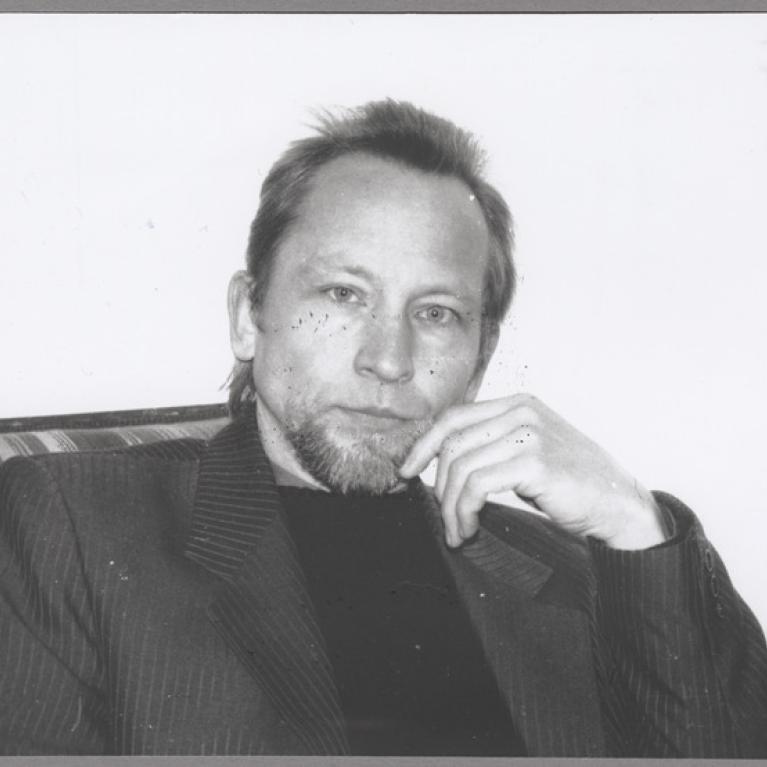 Black and white photograph of a person sitting on a striped couch, wearing a dark pinstripe jacket and a black turtleneck. The person is resting their head on their hand with an indoor plain background and a small portion of a table visible on the right.