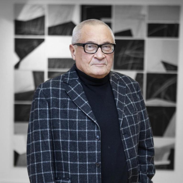 Photograph of a man with short grey hair wearing glasses in a checkered blazer against the backdrop of an artwork in the exhibition space