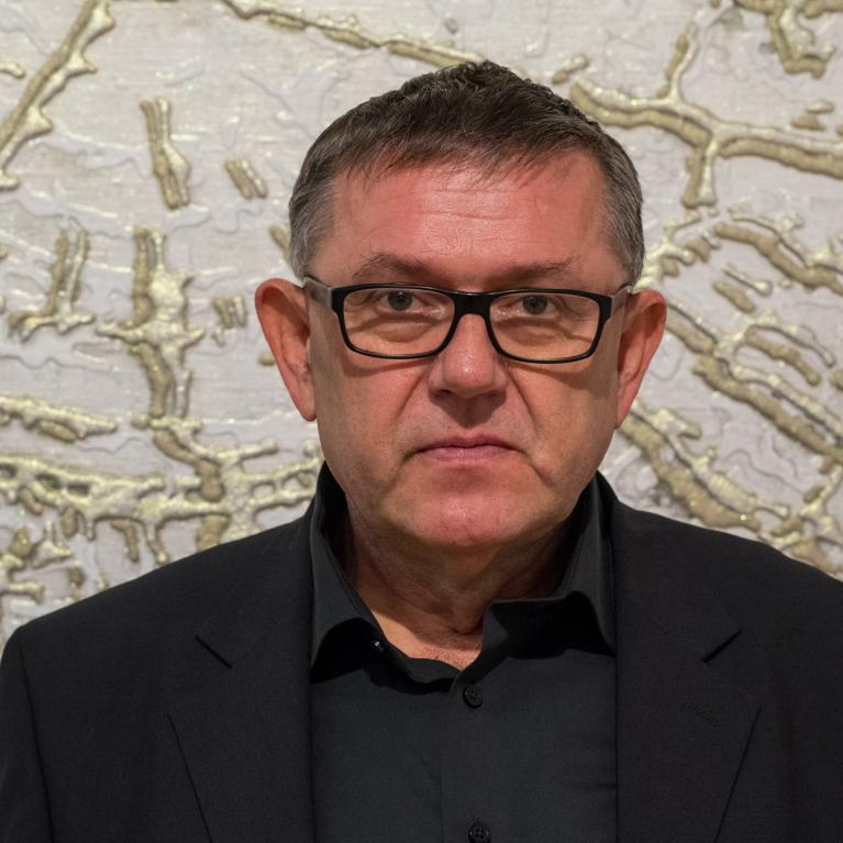 middle-aged man wearing glasses and a formal suit at the backdrop of a wall with golden pattern