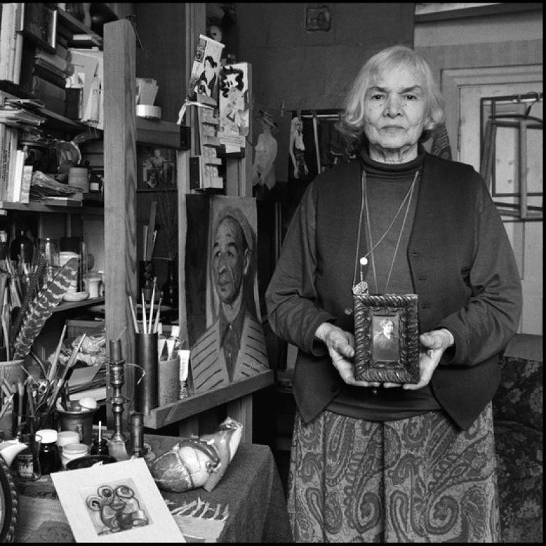 grey-hair woman of late age standing in art studio holding a small painting