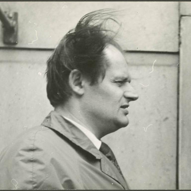 a man in a trench in front of a stone building, his hair flying in the wind