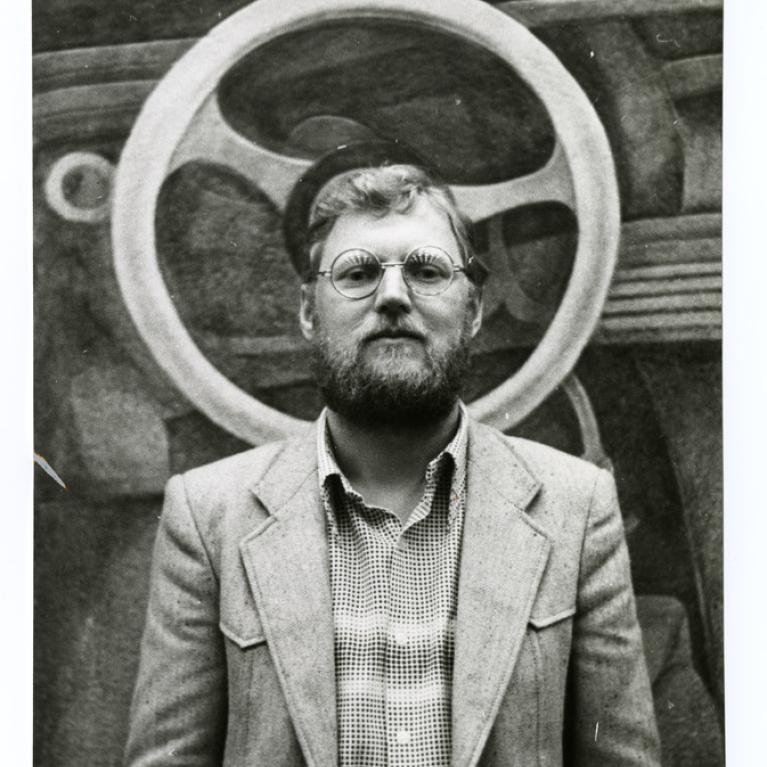 a young blonde man with beard and glasses with a painting of a wheel behind his head