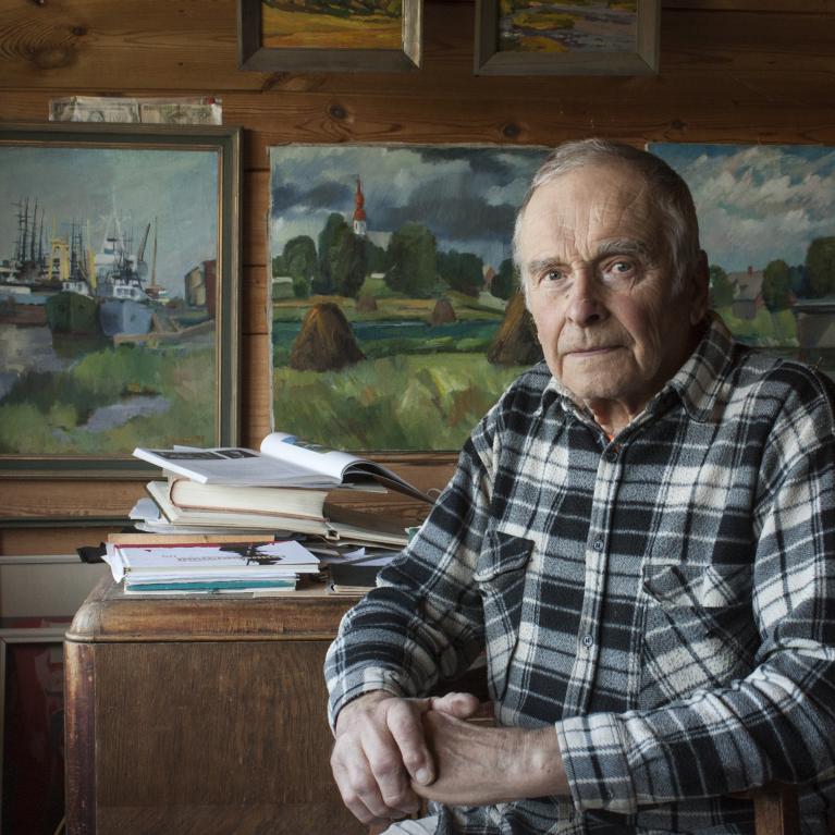 older man in checkered shirt, seated holding his hands; landscape paintings and books in the background