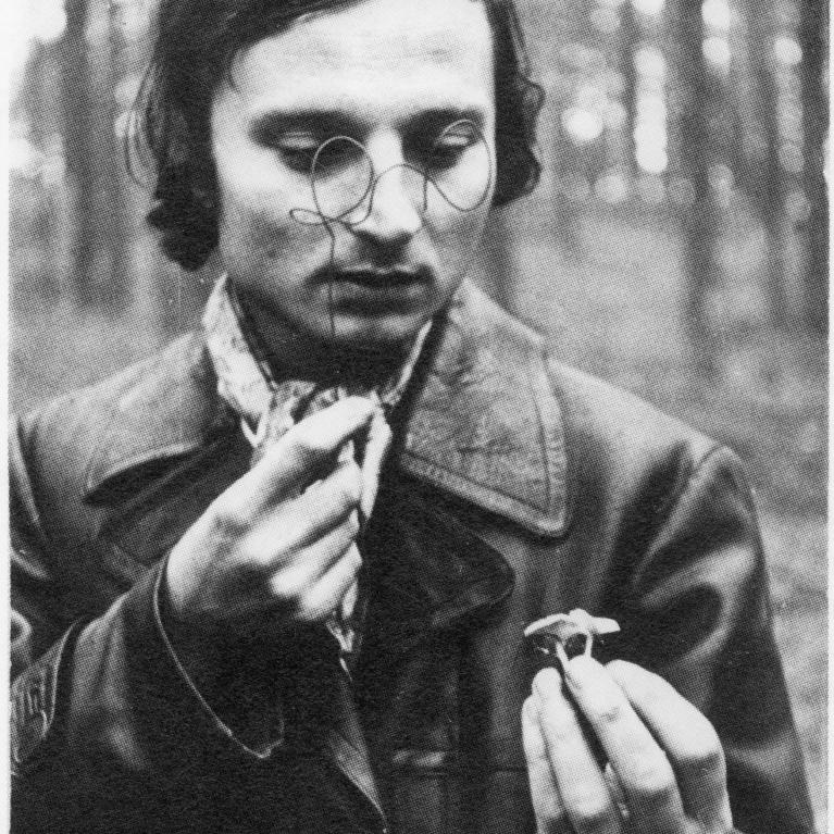 a young man in leather coat in the forest looking at a flower-shaped ring that he holds in his hand through a wire shaped as glasses that he holds in another hand