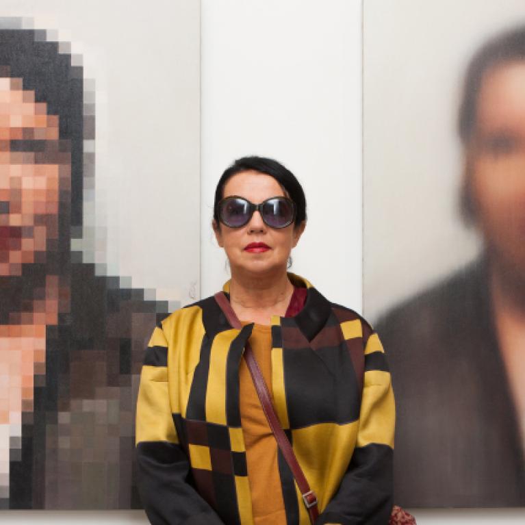 middle-aged woman in sunglasses standing against the background of two large-scale pixelated portraits of herself