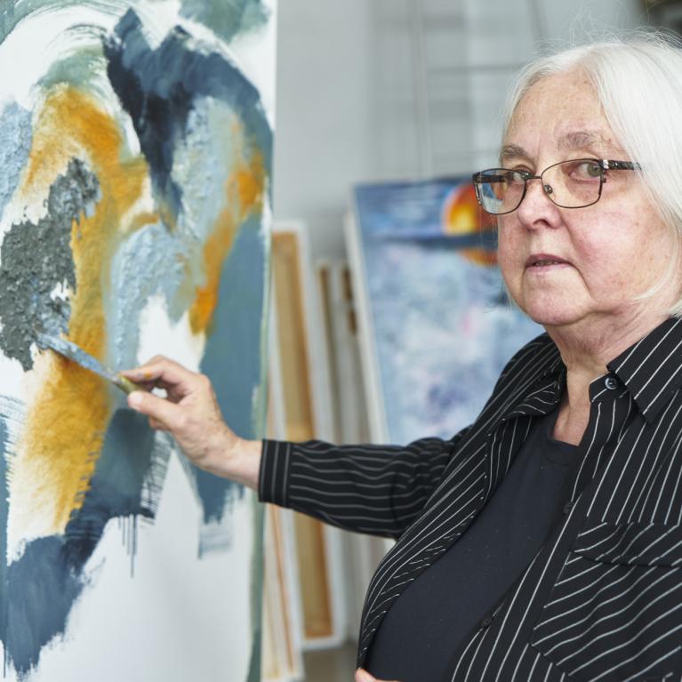 a woman with grey long bob wearing glasses standing in front of a painting with a paint brush