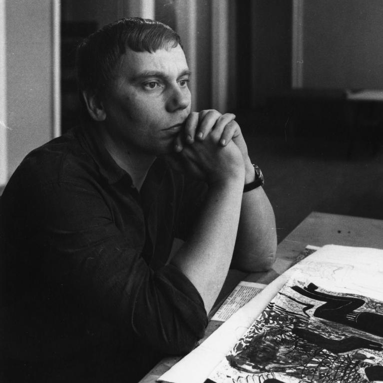 a man sitting at the table with artworks on paper on it, his hands under his chin, looking into the distance