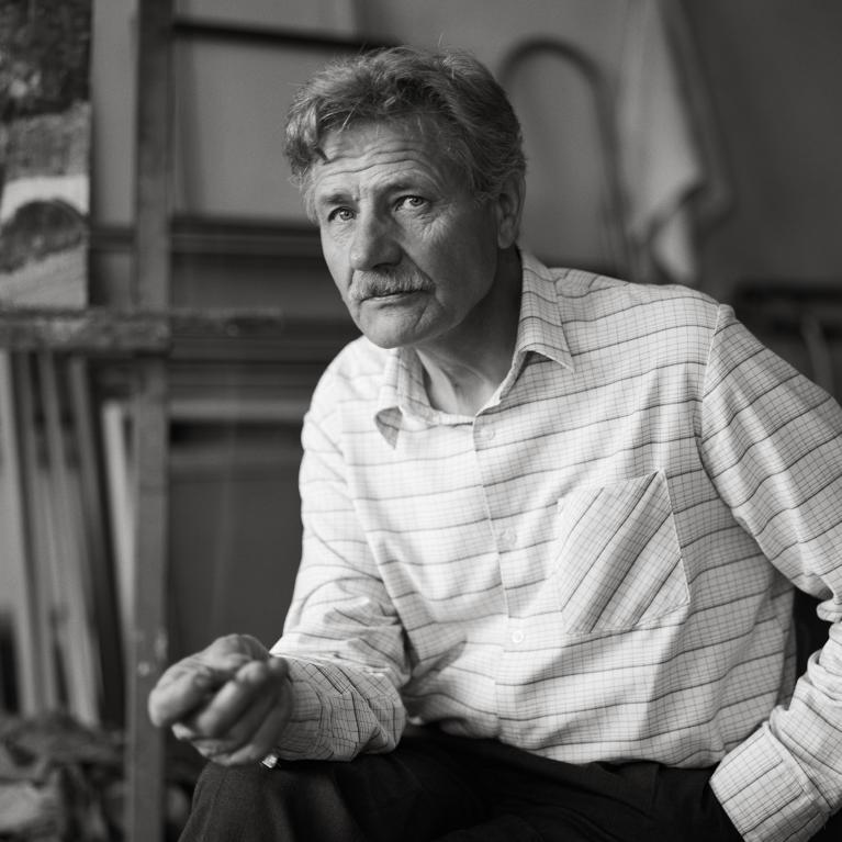 Black and white photograph of a man seated indoors, wearing a striped button-up shirt and dark pants. Background includes shelves with canvases and a hanging cloth