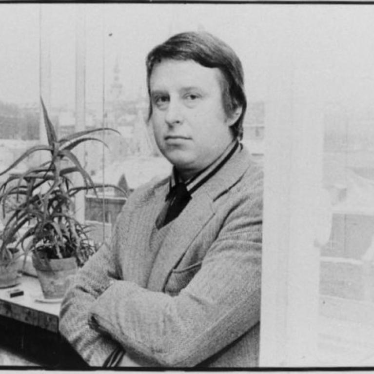 Black and white photograph of a person standing indoors near a window with arms crossed, wearing a sweater over a collared shirt. Several potted plants are arranged on a windowsill, and an outdoor scene with indistinct structures is visible through the window