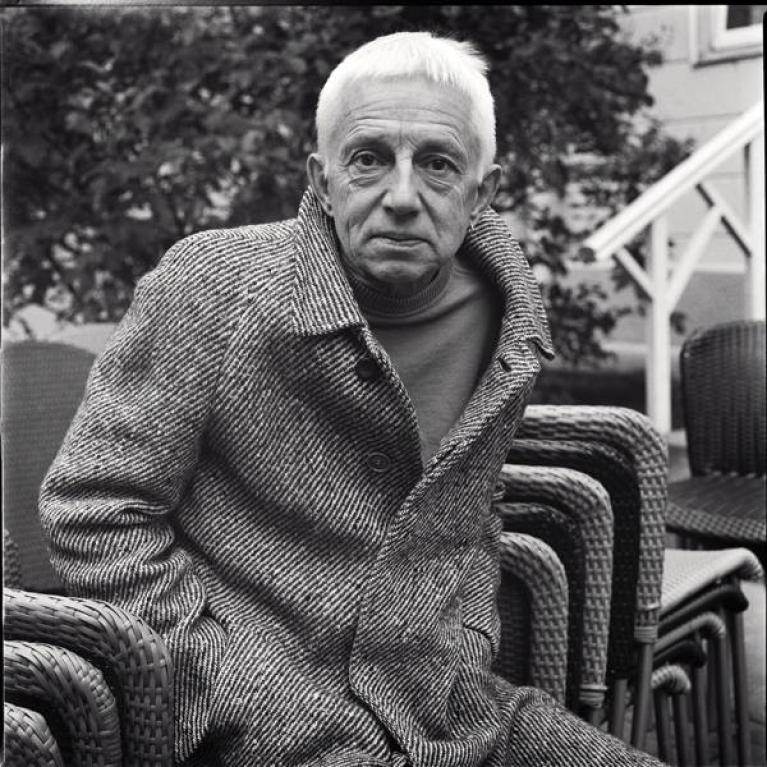 Black and white photograph of an elderly person wearing a textured coat sitting outdoors on a woven chair with several similar chairs stacked behind. Background includes foliage and part of a building with stairs, suggesting a garden or patio setting.