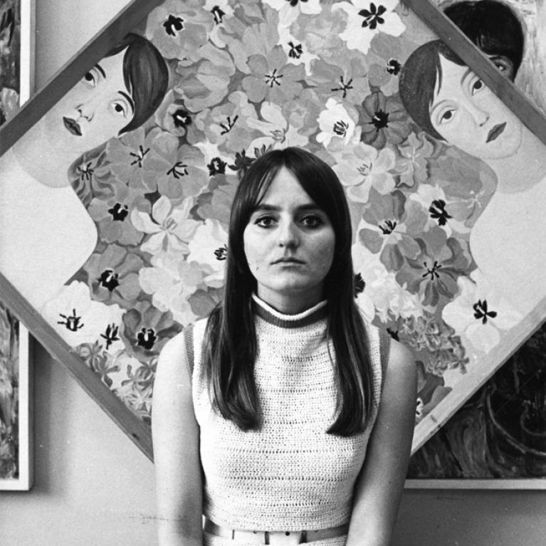 Photograph of a young woman in knit top with long hair and fringe seated in front of a colorful painting featuring multiple stylized female faces surrounded by floral patterns arranged in a diamond shape.