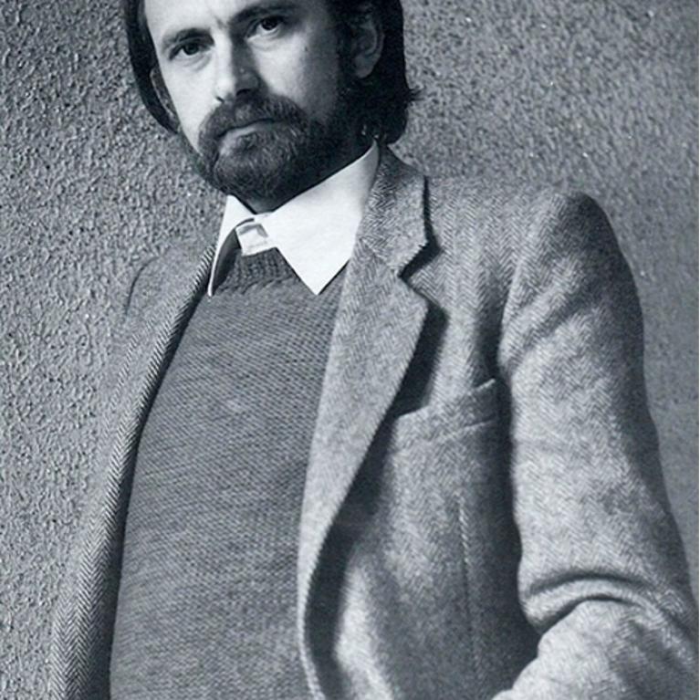 Photograph of a bearded man wearing a textured blazer over a sweater and collared shirt, standing against a rough, speckled wall.