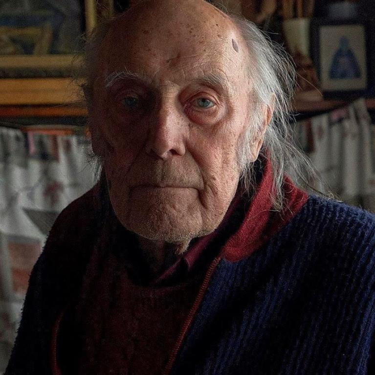 Photograph of an elderly man with gray hair wearing a dark blue sweater and a red shirt. Background includes framed artwork and patterned curtains.