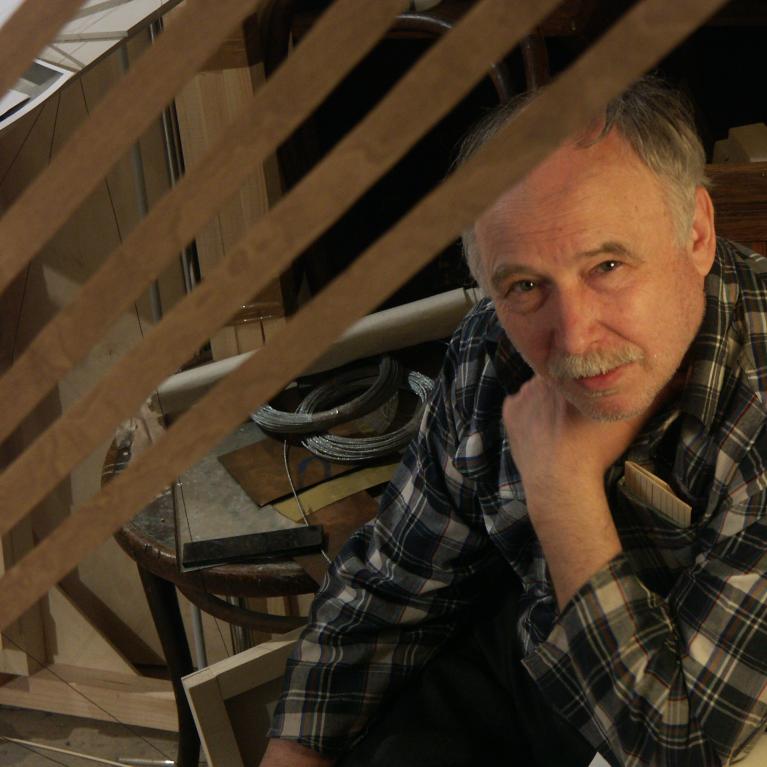 Photograph of a person wearing a black and white plaid shirt and a grey moustache sitting indoors near wooden structures and various tools. The setting suggests a workshop or construction area with visible wood beams, coiled cables, and a workbench in the background