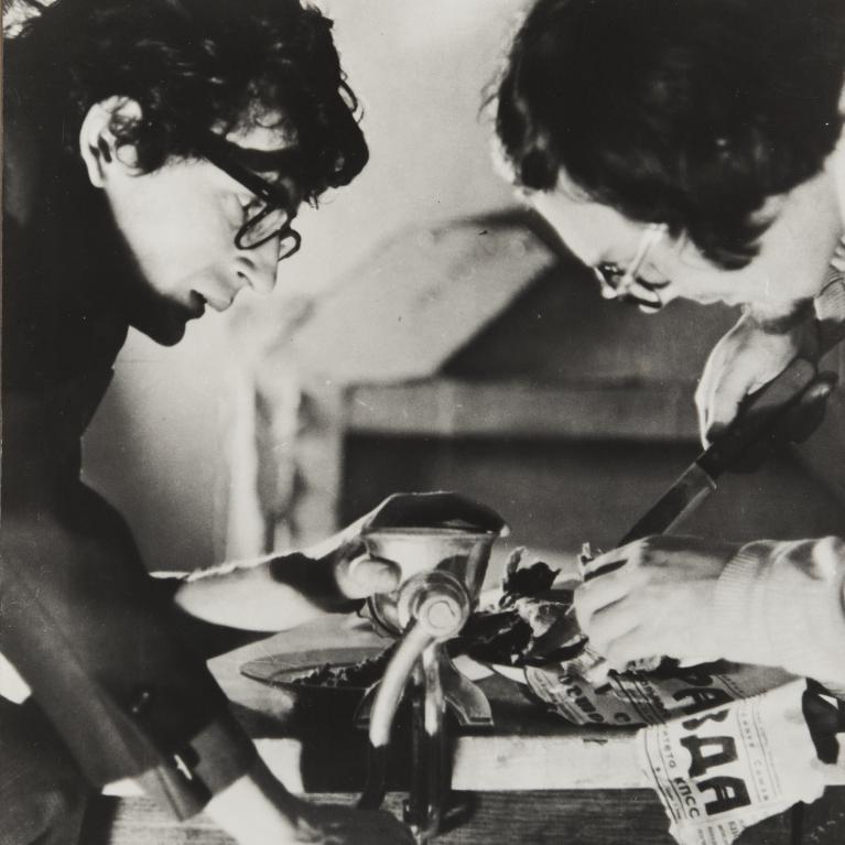 Black and white photograph showing two individuals working together on a manual task involving a grinder and a knife. One person holds a grinder while the other cuts newspaper, highlighting a collaborative effort in a workshop setting.