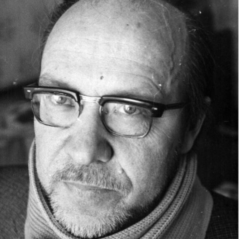 A black and white photograph showing an elderly man wearing glasses and a knitted scarf. 