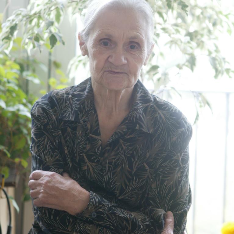 Photograph of an elderly person sitting indoors near a window with natural light and green plants in the background. The person is wearing a dark shirt with a leafy pattern and has their arms crossed gently across their chest.