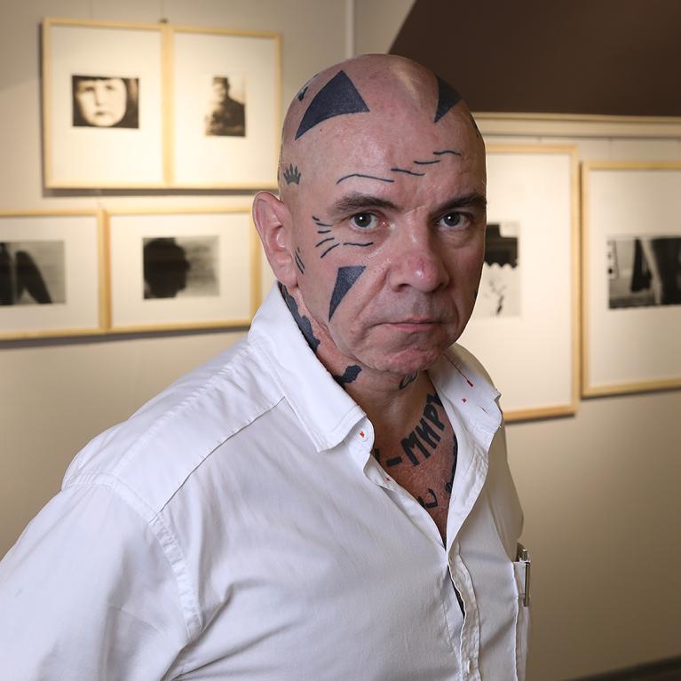 Photograph of a person with tattoos on their head and neck, wearing a white button-up shirt, standing in front of a wall displaying framed black-and-white photographs.