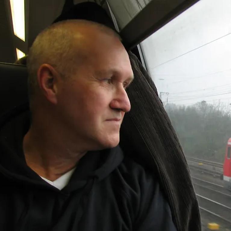 Photograph of a person wearing a dark hoodie sitting inside a train, looking out the window. Outside the window, railway tracks and a red train carriage are visible on a foggy day.