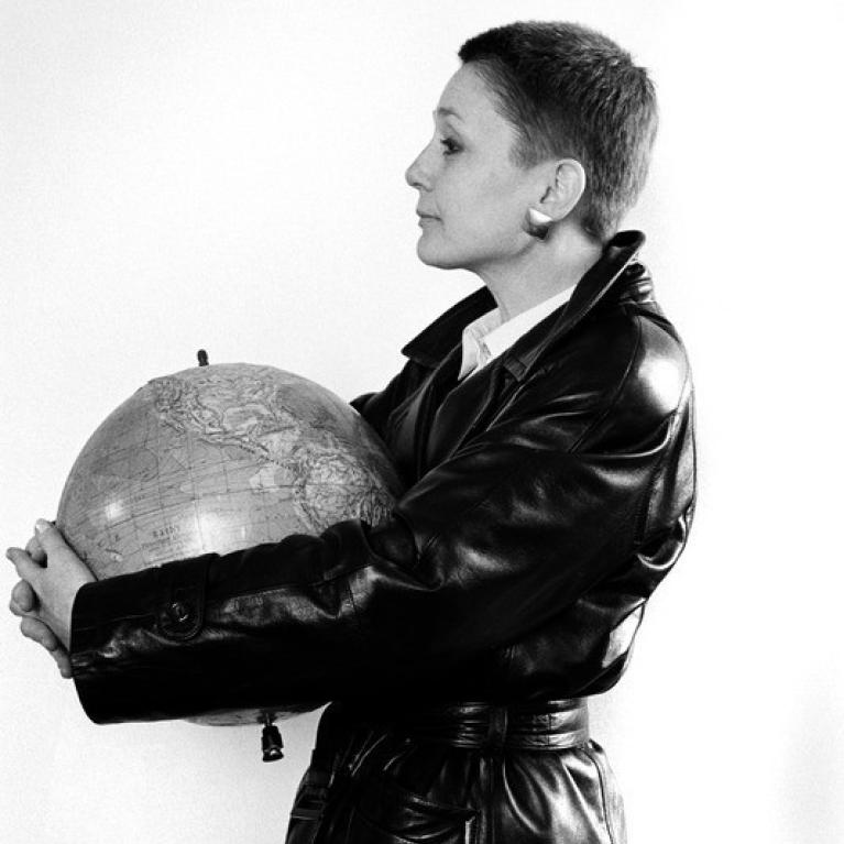 Photograph of a woman with short hair wearing a black leather jacket holding a globe close to their chest while facing sideways. 