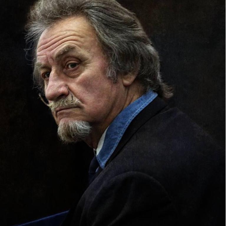 Photograph of a man with gray hair and Salvador-Dali-like moustache wearing a dark blazer and blue shirt, against a dark background. 