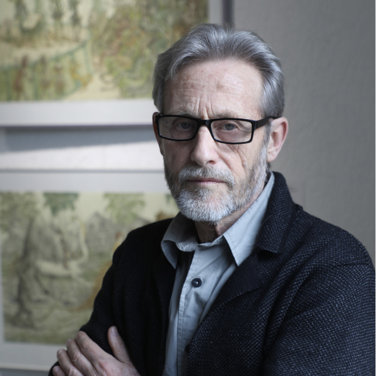 Photograph of an older man with gray hair wearing a dark textured jacket and light-colored shirt, standing with arms crossed in front of two framed landscape paintings. 