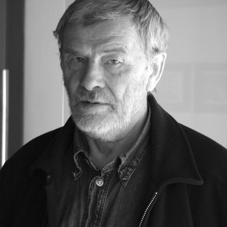 Black and white photograph of a man wearing a dark jacket over a buttoned shirt, standing indoors against a plain background. 