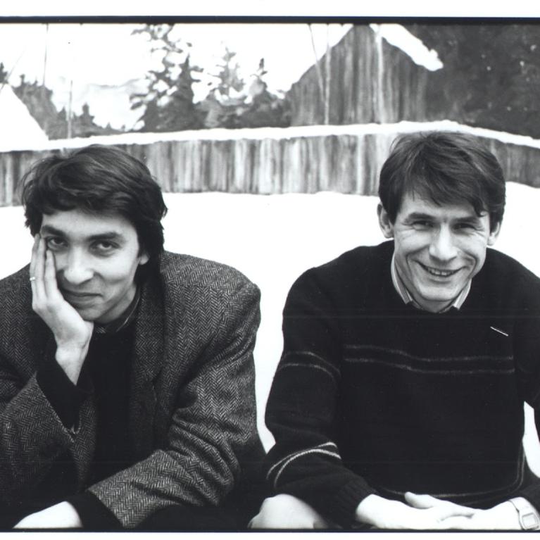Black and white photograph featuring two individuals sitting outdoors in a snowy setting with wooden structures and trees in the background. Both wear winter clothing, one in a textured coat and the other in a striped sweater, with relaxed postures suggesting a casual moment.