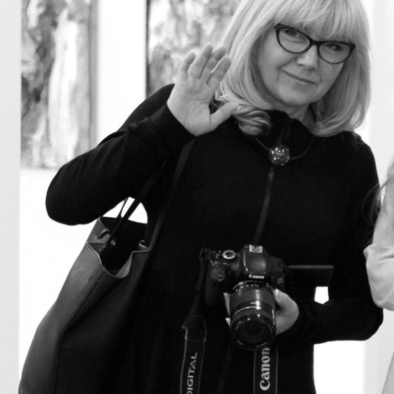 Black and white photograph of a woman with blonde straight hair wearing glasses and a dark outfit, holding a Canon camera with a visible strap, and carrying a large handbag and making a waving gesture with one hand