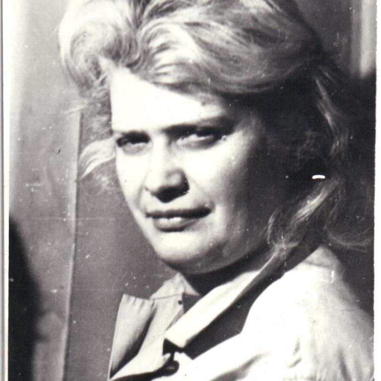 Black and white photograph of a person with voluminous, styled hair wearing a light-colored coat or jacket. The background appears plain, focusing attention on the individual's upper body and hairstyle.