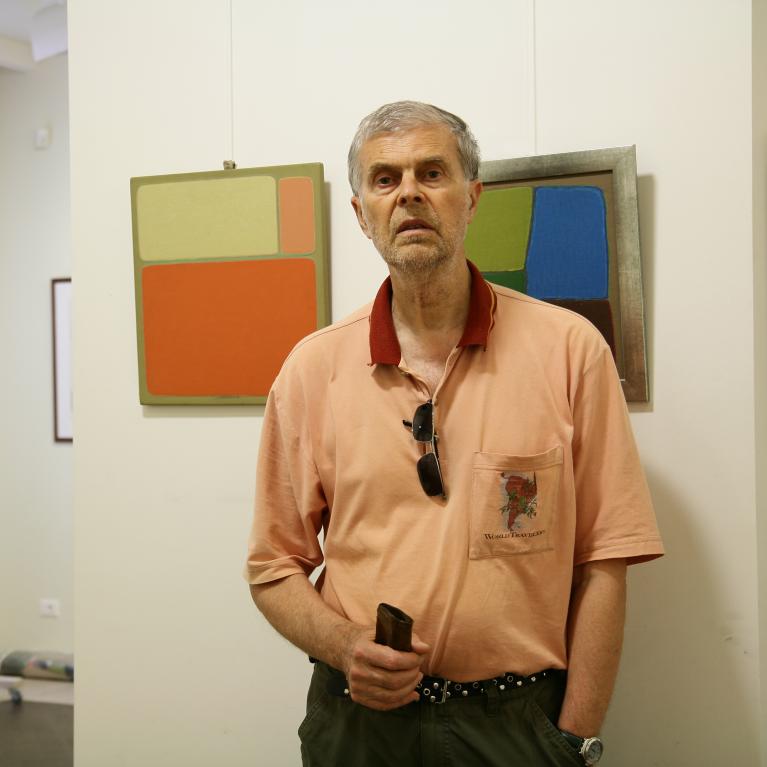 Photograph of a man standing indoors in front of a white wall displaying two abstract paintings with geometric shapes in muted colors like orange, green, and blue. The man wears a peach-colored polo shirt with a small graphic on the pocket, sunglasses hanging from the collar, and holds something in his right hand.